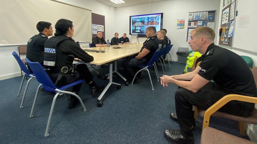 Officers being briefed as part of the operation in a meeting room at the police station. Some are sitting at a table, while others are sat on chairs away from it. There are various posters and leaflets on the walls and on cork boards. There is a computer monitor on and it appears to feature officers at other places on a live meeting.