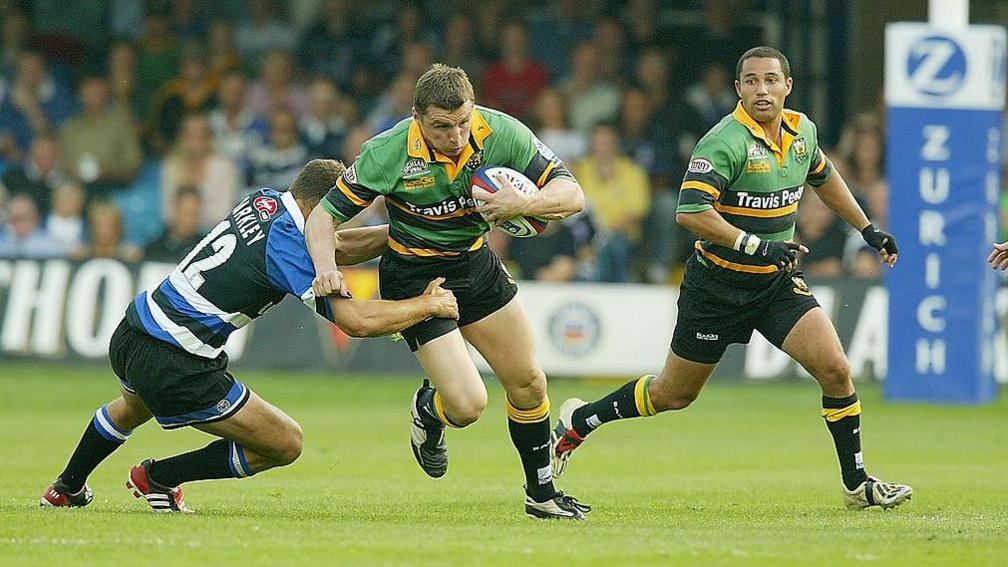 A man in a green and yellow rugby shirt carrying a rugby ball runs past a man trying to tackle him. 