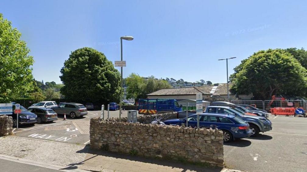 Mayors Avenue car park on a sunny day is mostly full of parked cars. A stone wall separates the car park from the road and there are trees lining the left hand side and back of the area. There are street lamps in the car park and indecipherable signage.