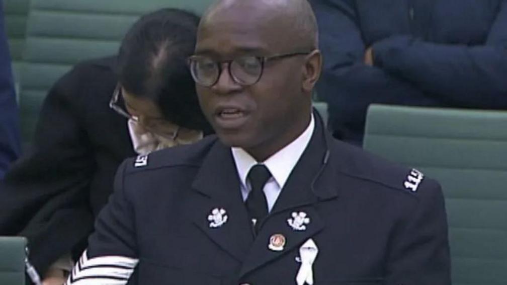 Inspector Tola Munro wearing his police uniform and glasses while sitting on green chairs in a committee meeting.