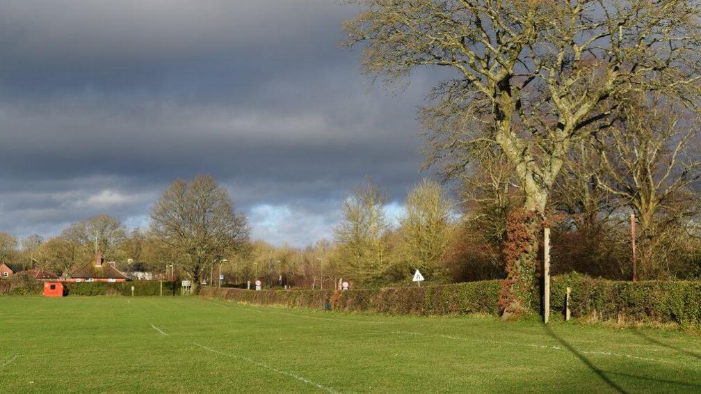 View along the edge of the recreation ground. A playing field is in the foreground, with trees and a hedge to the right.