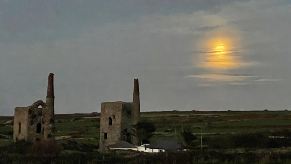 This image depicts a rural landscape at twilight or early evening, featuring two old stone mining structures, surrounding green fields and low vegetation, and a bright full moon in the background, partially veiled by clouds.
