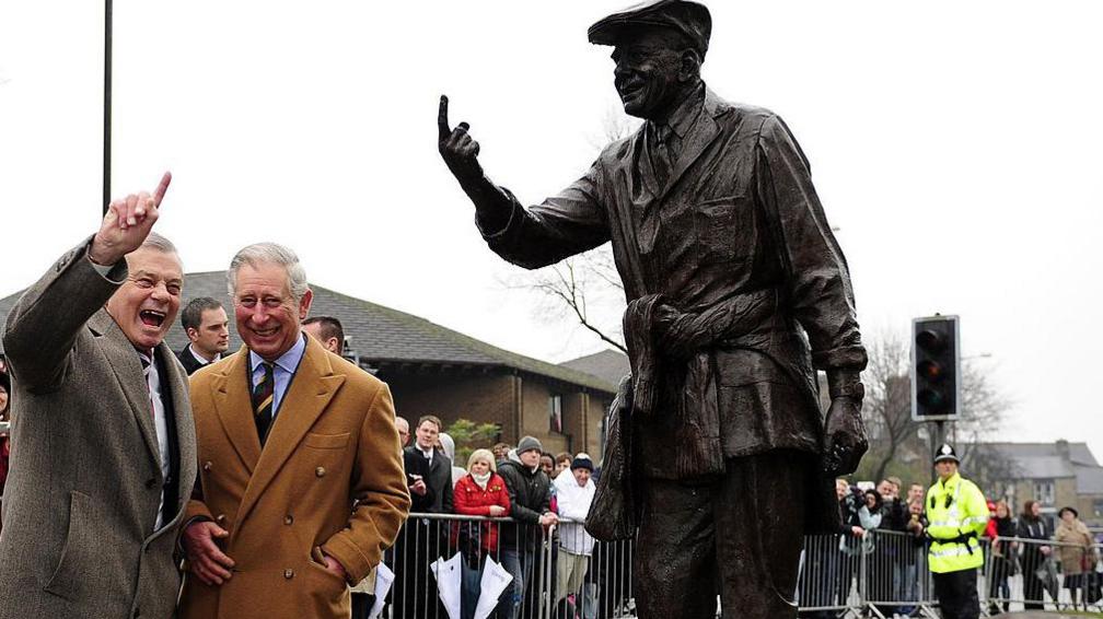 The then Prince of Wales, wearing a camel overcoat, stands between former cricket umpire Dickie Bird as they both look at Bird's statue in Barnsley