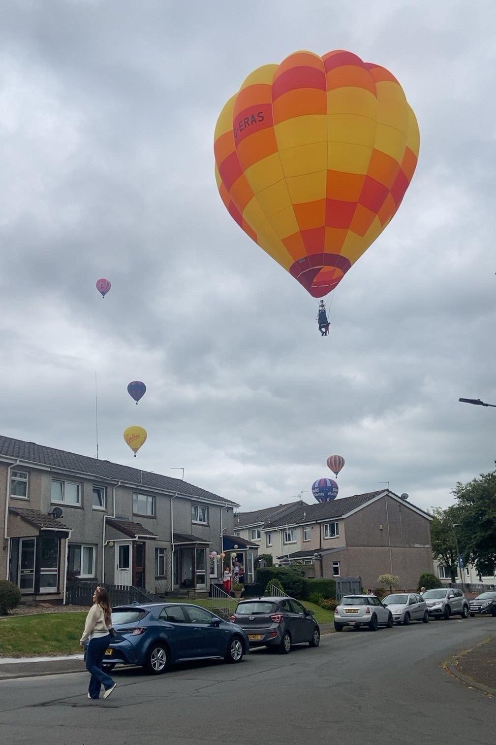 Hot air balloons of different colours hang in the sky above a street.