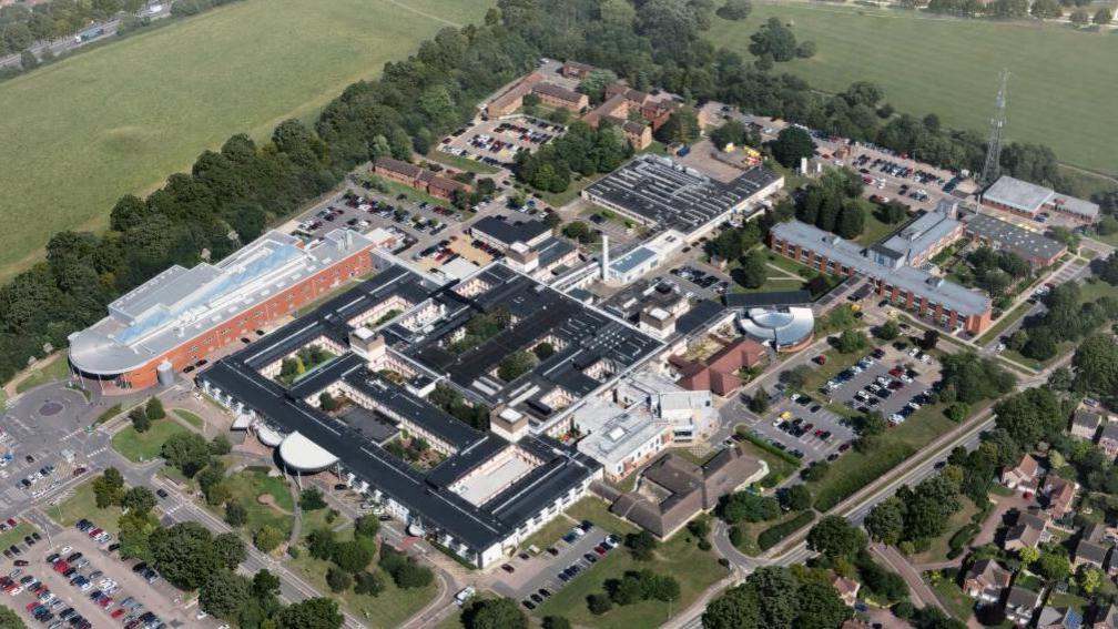 An aerial view of Hinchingbrooke Hospital, which is a sprawling campus covered in large buildings. It is surrounded by fields, trees and some houses.