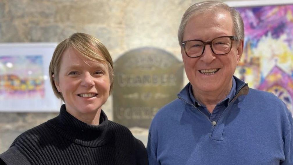 Alice Gill and Peter Neville are smiling while standing next to each other. Alice Gill has blonde bobbed hair and is wearing a black roll neck jumper. Peter Neville is wearing glasses and a blue shirt with a blue sweatshirt on top. A blurred sign saying Guernsey Chamber of Commerce can be seen behind them.