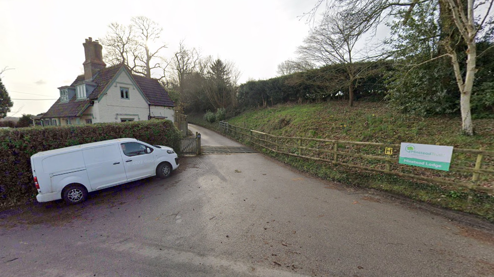 A lane with a cattle grid leading to a cream two-storey house. There is a white van parked at the entrance to the lane. A green sign on a fence reads: "Minstead Trust" and "Minstead Lodge"