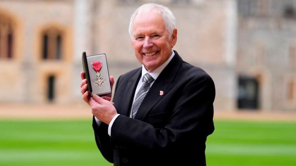 Nick Owen holding his MBE medal in an open case. The medal is silver with a red ribbon. Owen is wearing a dark suit, white shirt and silver striped tie. Behind him is Windsor Castle, which is out of focus, and a green lawn. 