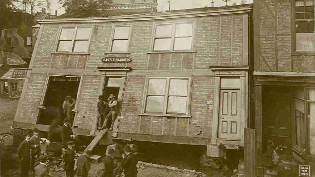 A building called Castle Chambers has sunk backwards, leaving a hole in the ground beneath it. A number of men are around it, with three men standing in the doorways of the building looking out. 