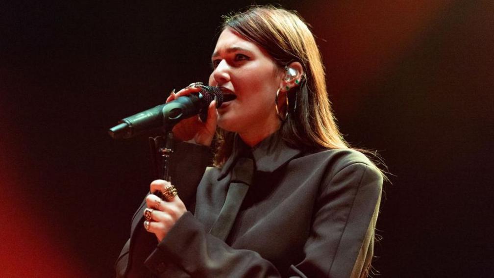 A young woman sings into a microphone. She's wearing an all-black suit with matching tie. Her long, straight brown hair spills down her back and she wears a selection of gold jewellery - multiple rings, hooped earrings and studs.