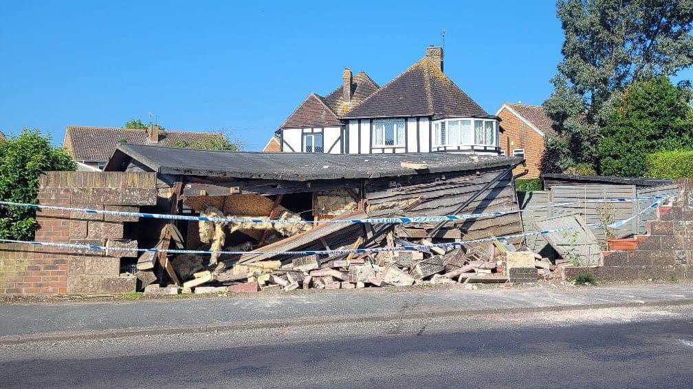 Crumbled bricks on the floor on the same section of wall from 1 June last year. Police tape is stretching across between the two sections of wall which are still standing. The garden shed is also damaged with one of the walls caved in. Behind there is a large white house which is next to Mrs Vilday's house. The skies are blue and clear 