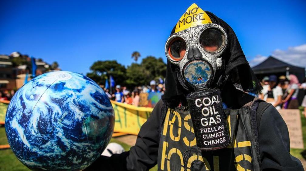 A climate protester wearing a gas mask and protective clothing holds a model of planet earth. Thier face his obscured bythe mask that reads in captial letters: NO MORE COAL OIL GAS FUELLING CLIMATE CHANGE. 