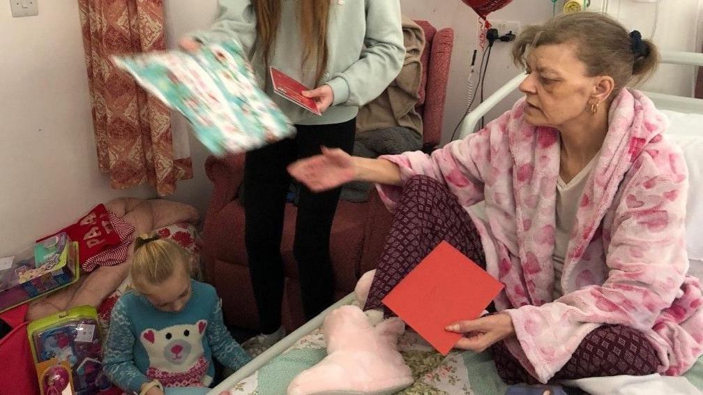 Carol sitting on her hospice bed in fluffy pink slipper-boots, a fluffy pale pink dressing gown with hearts on, and pyjamas underneath which are purple (trousers) and white (vest top). Her hair is tied back into a ponytail and she had gold hoop earrings. She holds a red envelope in one hand and is reaching out to give or take a present. A young girl sits on the floor reading a card and another girl, who can't be seen in full, holds a big piece of wrapping paper.