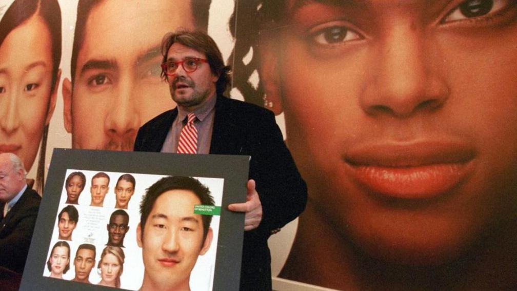 Toscani wears a dark jacket and holds a display of models of different ethnicities he has photographed. He has long hair, and wears a red and white striped tie and red framed glasses. 
