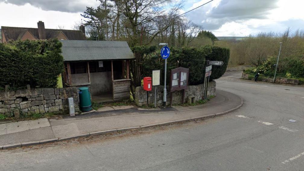 A street view image of a small red post box at the side of a road, next to a junction. An old wooden bus shelter is to its left with a notice board and various road signs to the right.