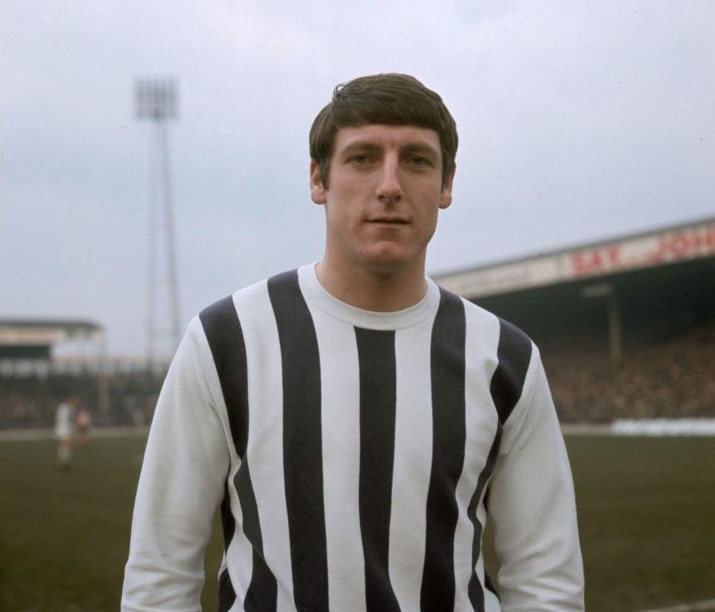 A man with dark hair is wearing a striped football top. He is standing on a football pitch with two stands at a football ground and a large floodlight pylon visible in the background.