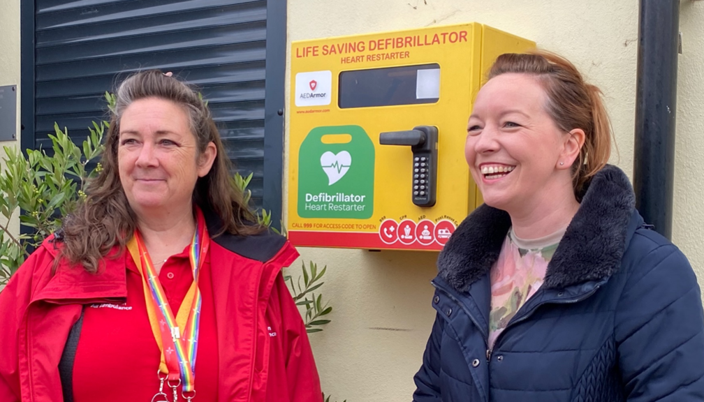 Simone Black and Claire Whitehouse standing beside a defibrillator box on a wall