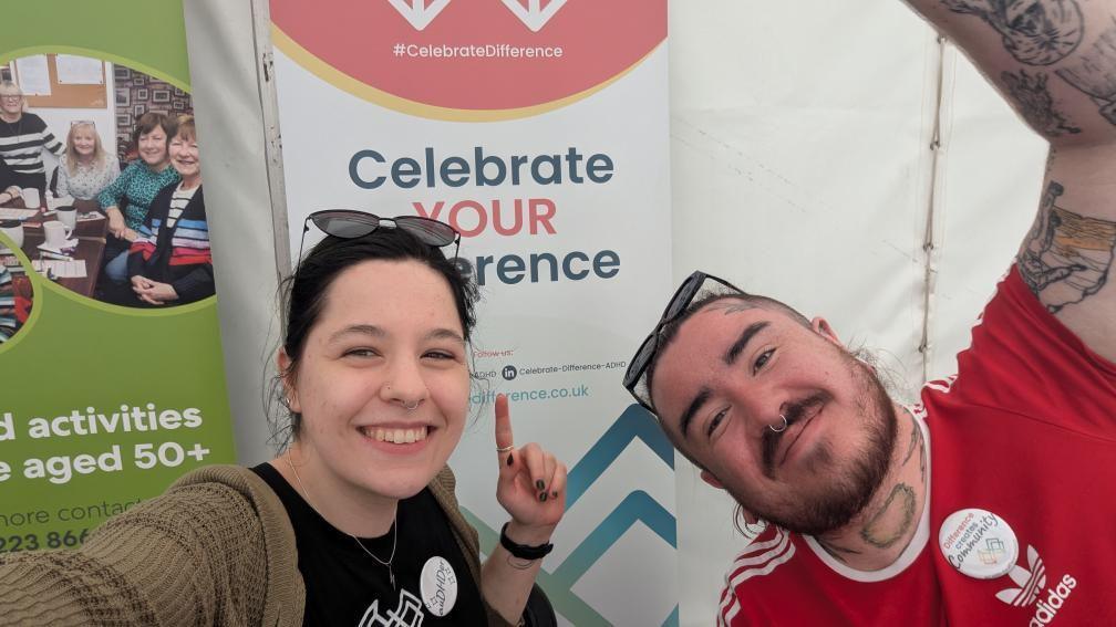 Holly Knight smiling and pointing to a banner behind her reading Celebrate Your Difference. She is wearing a black t-shirt and a beige cardigan with sunglasses on top of her head. Her black hair is tied back. Her colleague Michael is smiling next to her in a red t-shirt. He also has sunglasses and a nose ring. They are at the Hub Consett stall.
