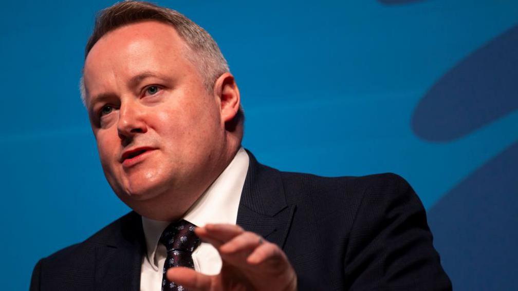 Darren Millar speaks at previous conference in navy suit and white shirt with blue background and gestures with one hand 