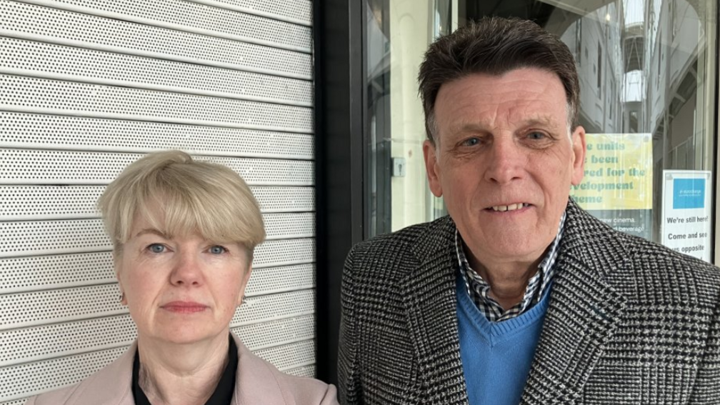 A man and a woman stand in front of shops with shutters down. The woman has blonde hair and is wearing a pink coat. The man has dark brown hair and is wearing a blue jumper under a coat made from black and white houndstooth material.