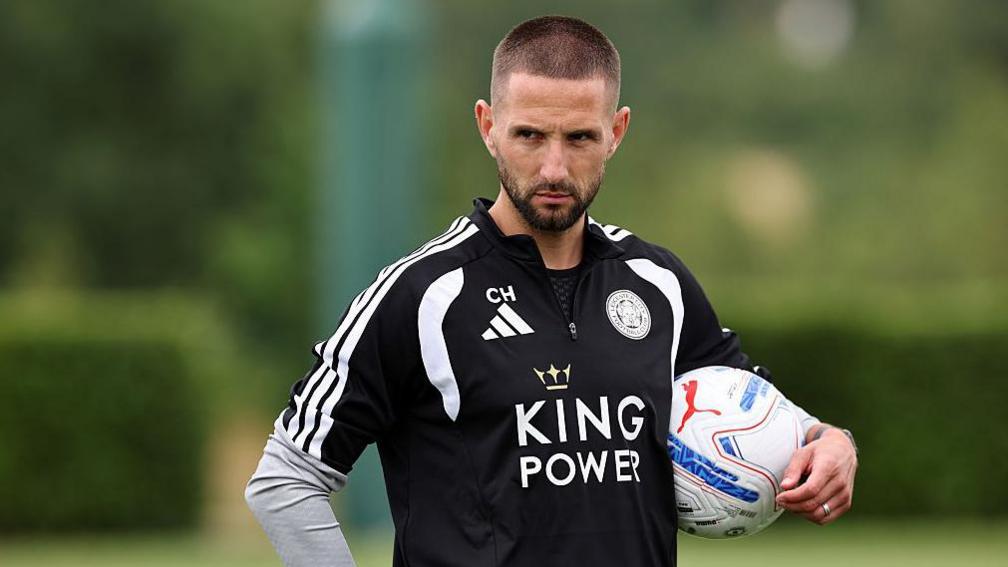 Conor Hourihane wearing a black Leicester City training top with King Power written across the middle and holding a football under his arm while watching training