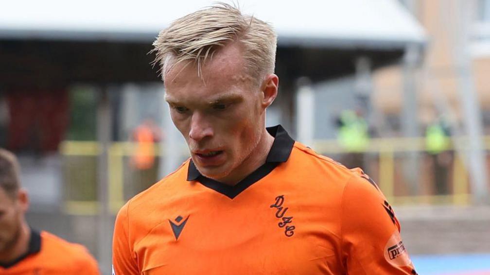 Ilmari Niskanen wearing an orange football shirt from his time at Dundee United. He is looking down at the floor. There are other Dundee United players out of focus behind him. 