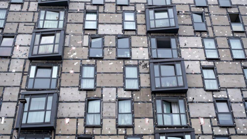 Image showing an apartment building which had its cladding removed in 2018 but was waiting for it to be replaced in July 2024. Brown boards and the internal struts and clips for the cladding are visible next to the black windows of the building