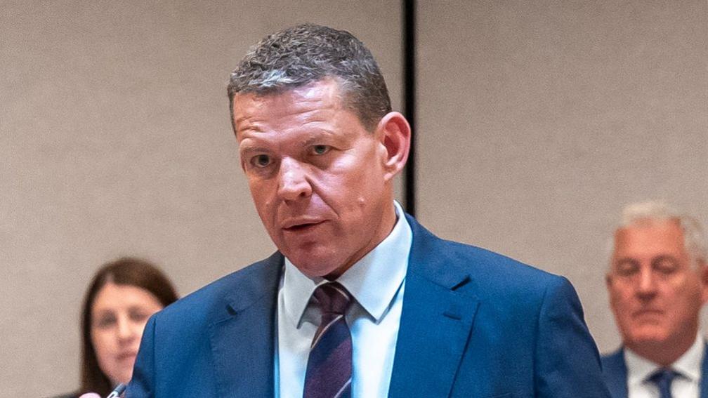 Rhun ap Iorwerth is wearing a blue suit, a light blue shirt and a red tie.