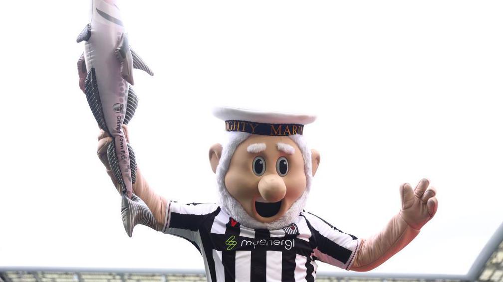 The Mighty Mariner, Grimsby Town's mascot posing for a photo with Harry Haddock - an inflatable fish. The mascot is wearing the team's black and white kit.