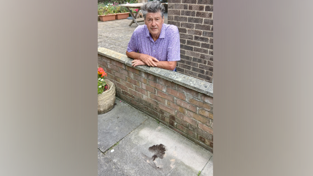 A black and brown marking in the shape of a tree on stone which looks like the Sycamore Gap tree. Howard Dawson, wearing a purple shirt, with long grey hair, poses next to it, leaning on a wall.
