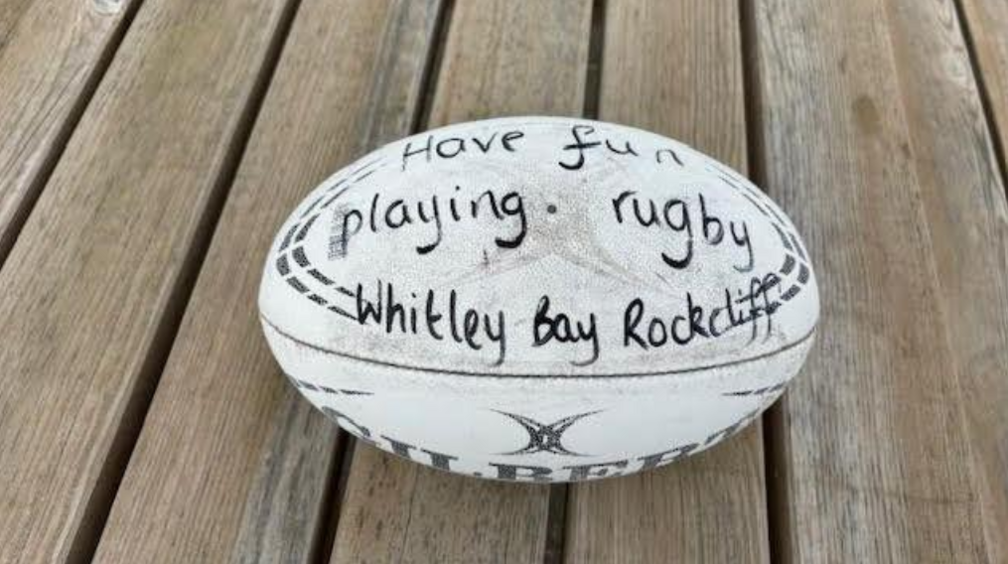 A rugby ball lying on wooden decking. The ball is a white Gilbert ball and appears well worn. A message has been written on in black market pen. It reads, Have fun playing rugby - Whitley Bay Rockcliff.