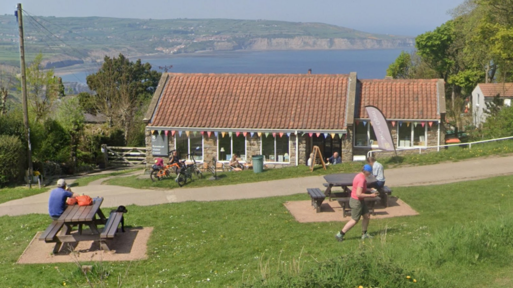 A small stone café with a red‑tiled roof sits on a grassy coastal hillside, decorated with colourful bunting, as people sit at picnic tables or walk and cycle nearby, with the sea and distant cliffs visible behind it.