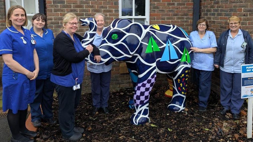 Six women in blue nursing uniforms gather around a large navy blue elephant decorated with white wave patterns and green and blue sail boats