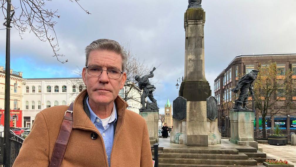 John Boyle stands in front of the war memorial in Derry. Mr Boyle is wearing a blue shirt, eye glasses, and a brown coat. The strap of a bag is over his right shoulder