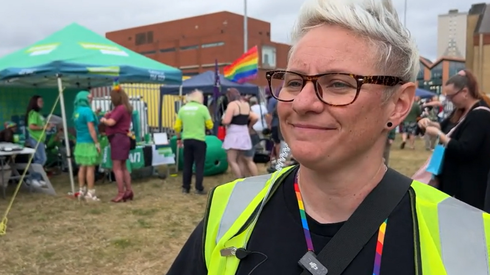 Event organiser Abi Pullin being interviewed at Pride in Scunthorpe, she is wearing a rainbow lanyard and a hi-vis vest. She has short, bleach blonde hair and tortoiseshell-rimmed glasses. 