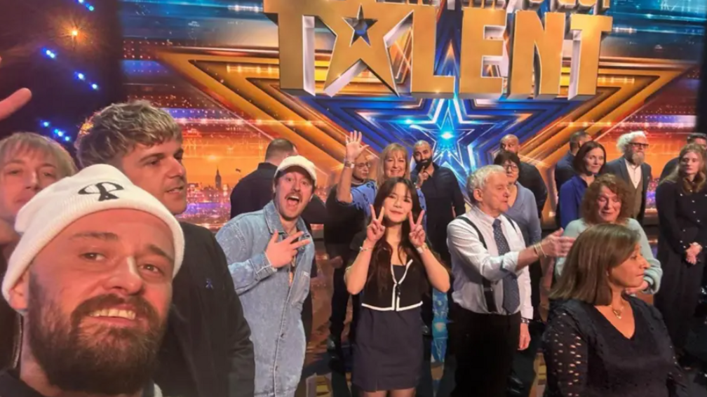 A bearded man in a white beanie hat standing alongside a group of people on a stage. The "Britain's Got Talent" branding is behind them.