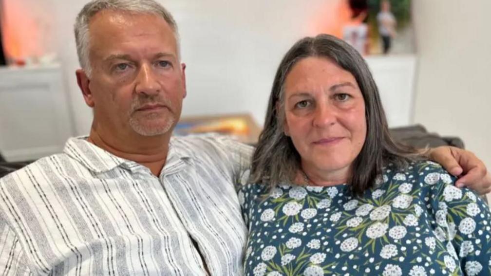 Middle aged man and woman sitting on a sofa. The man has his arm around the woman. He is wearing a white shirt. The woman has a floral top.