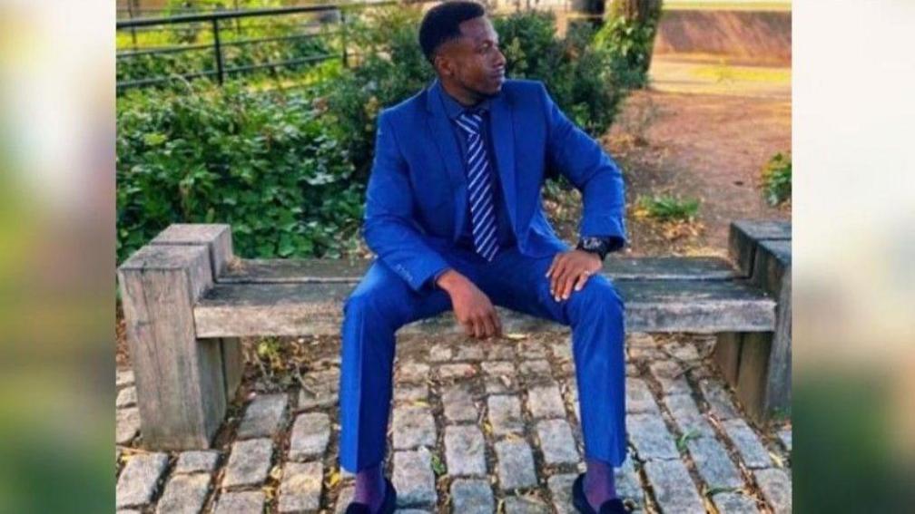 Allen Adeyemi Tejan is sitting on a bench looking to the right. He is wearing a blue suit and tie, black loafers, purple socks and a black watch. 