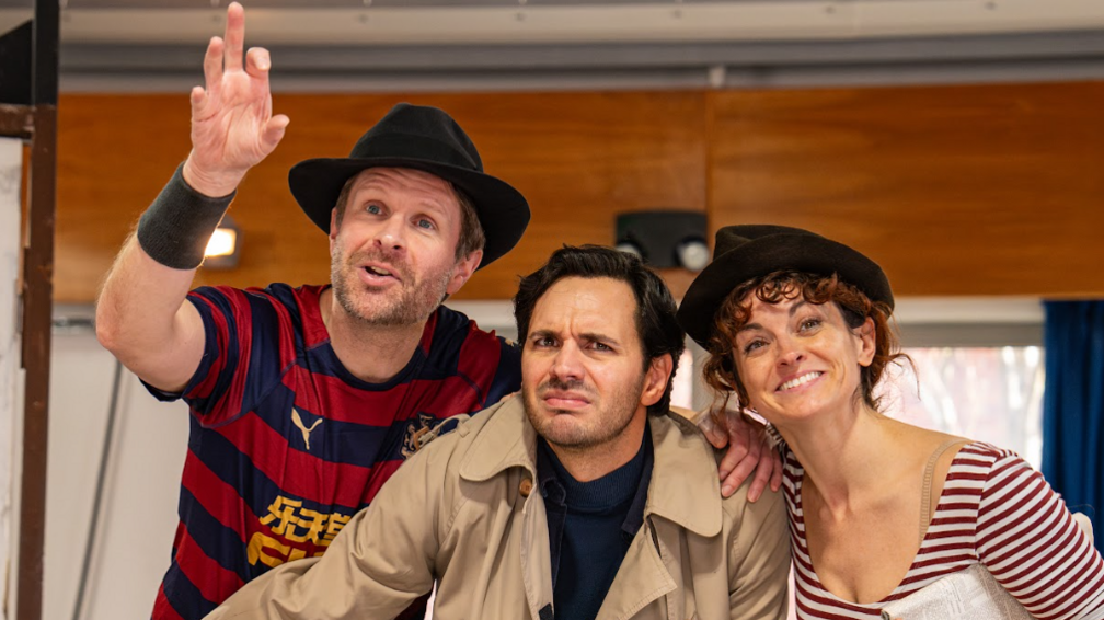 A close-up of three actors, one female, in rehearsal for The 39 Steps, two are wearing black trilbys and t-shirts, the other one is in a beige trench coat. 