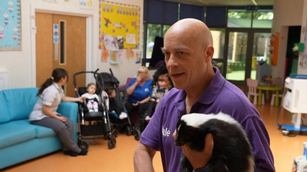 A bald man in a purple shirt holds a black and white skunk in his left arm while smiling. Behind him are several children in prams, with nurses next to them.