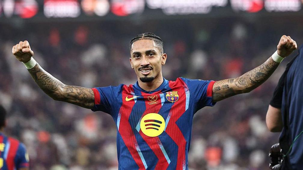 Raphinha, wearing a Barcelona shirt, spreads his arms wide in celebration