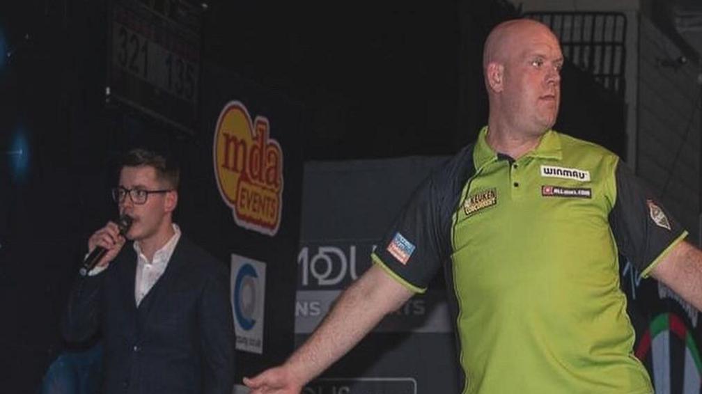 Owen Binks speaks into a microphone while wearing a white shirt and suit jacket on stage at a darts event. Michael van Gerwen, who is bald and wearing a green and black polo shirt, stands next to him with his arms spread.