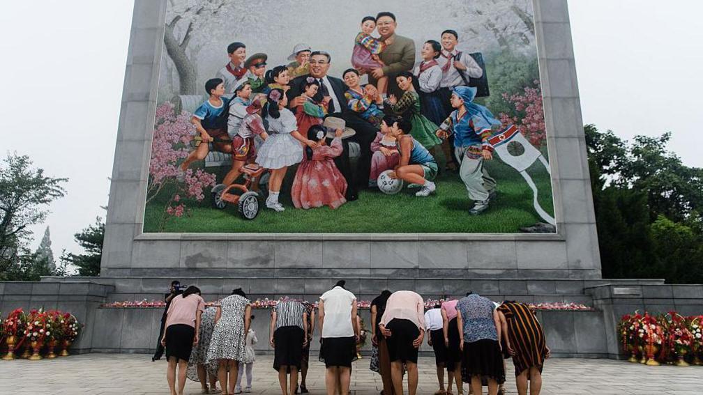 A group of women standing in two-three rows bow in front of a mosaic in Pyongyang. The mosaic shows Kim Jong Un's father and grandfather smiling as they are surrounded by beaming children in a garden, flanked by pink flower bushes. On their the side of mosaic, which sits on a stone square, are large bouquets of red and white flowers in gold-coloured urns. The photo was taken on the 77th founding anniversary of the Kim family's regime.