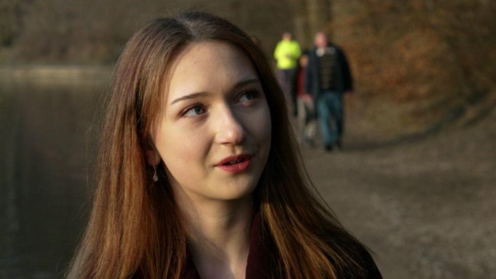 A close-up of a young woman with red hair who stands next to a lake. The outline of three people can be seen in the background, one of them wears a neon yellow top.