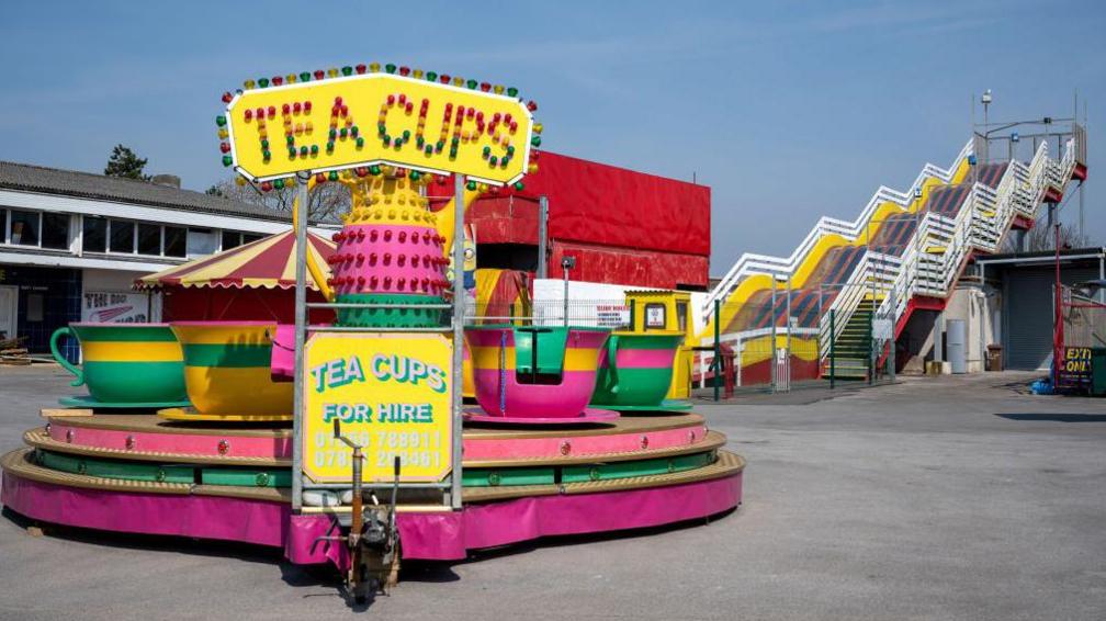 A teacups ride which is pink, yellow and green. Behind are the steps up to a large multi-coloured slide.