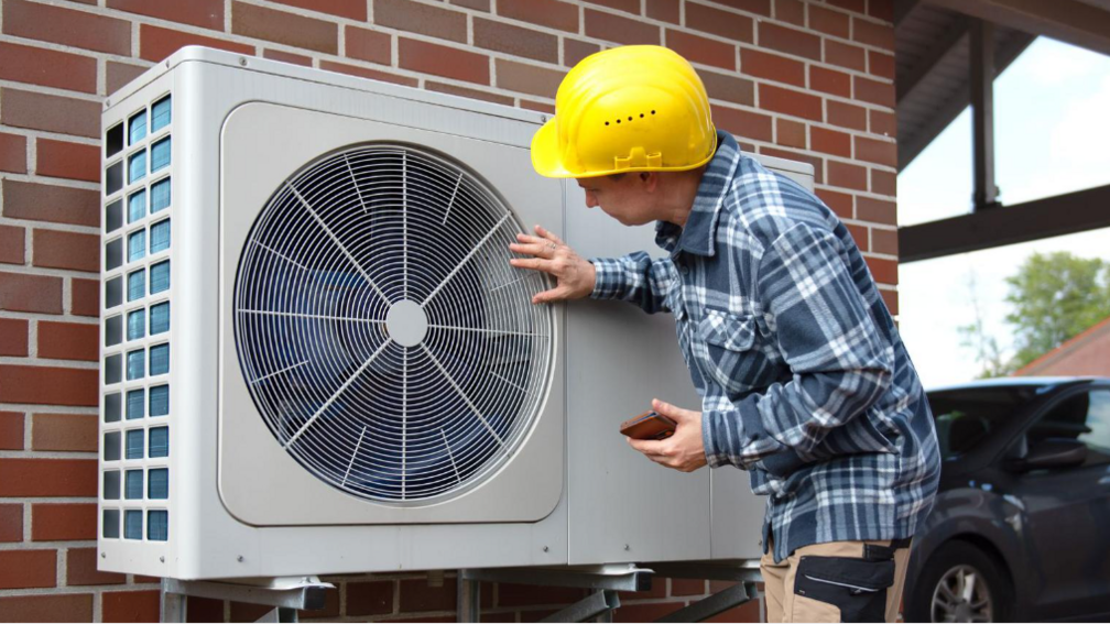 A man in a yellow hard hat examining an external heat pump on the wall of a house. The pump is a large grey box with a fan in it.