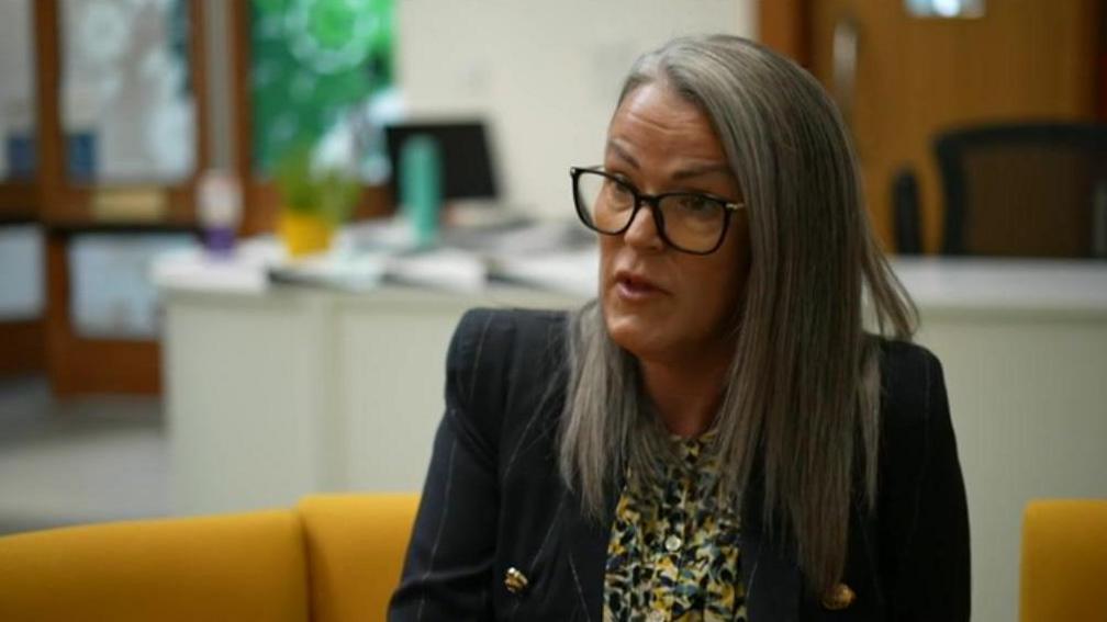 Helen Davis has long, grey and white hair past her shoulders and wears a black jacket over a multi-coloured blouse