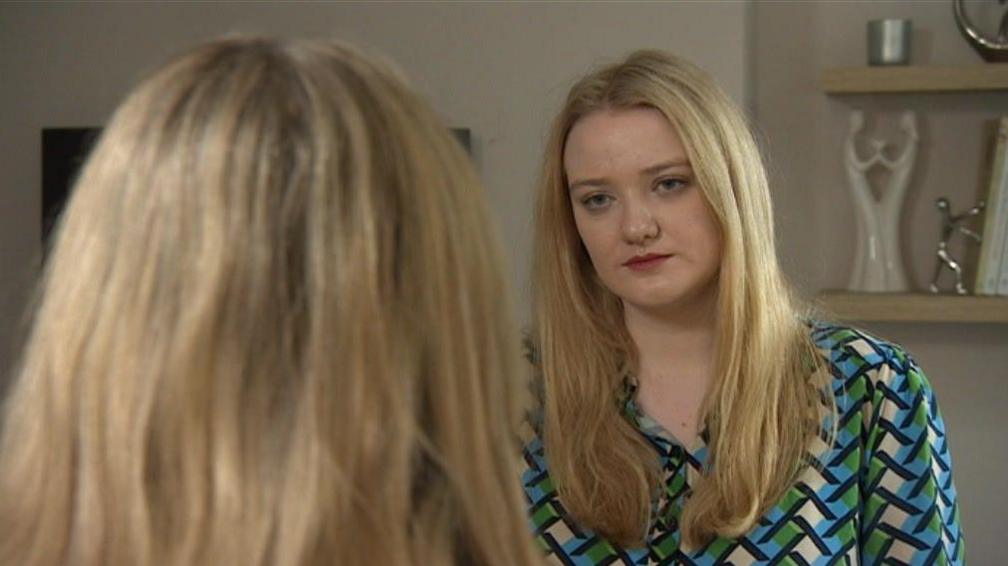 A BBC journalist with blonde hair wearing a blue and green patterned dress, looks at an interviewee who has the back to the camera. The interviewee is anonymous, so only the back of their head is visible. They also have blonde hair.