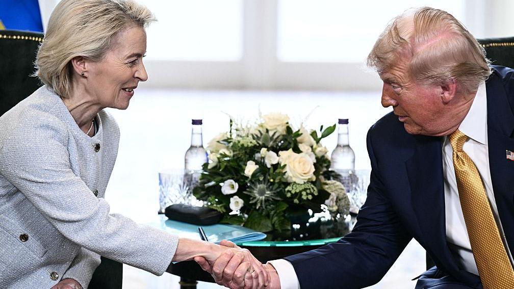Ursula von der Leyen shakes hands with Donald Trump, both seated with a bunch of white roses and Scottish thistles between them. Von der Leyen wears a light grey suit jacket and has short blonde hair. Trump wears a navy suit jacket, white shirt and a mustard tie.