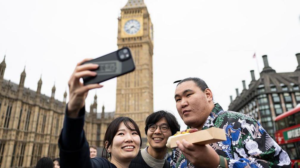 Sumo wrestler Yokozuna Hoshoryu, eating a hot dog, poses for a selfie with two people near Big Ben
